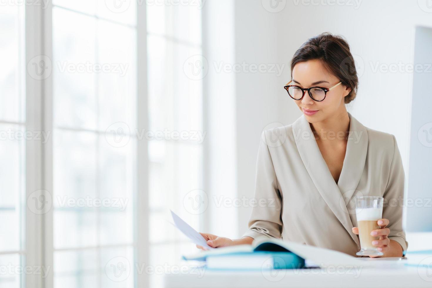 Woman economist checks information on business paper, wears spectacles and elegant suit, drinks milkshake, poses against office interior, being in cabinet, prepares startup project. Working process photo