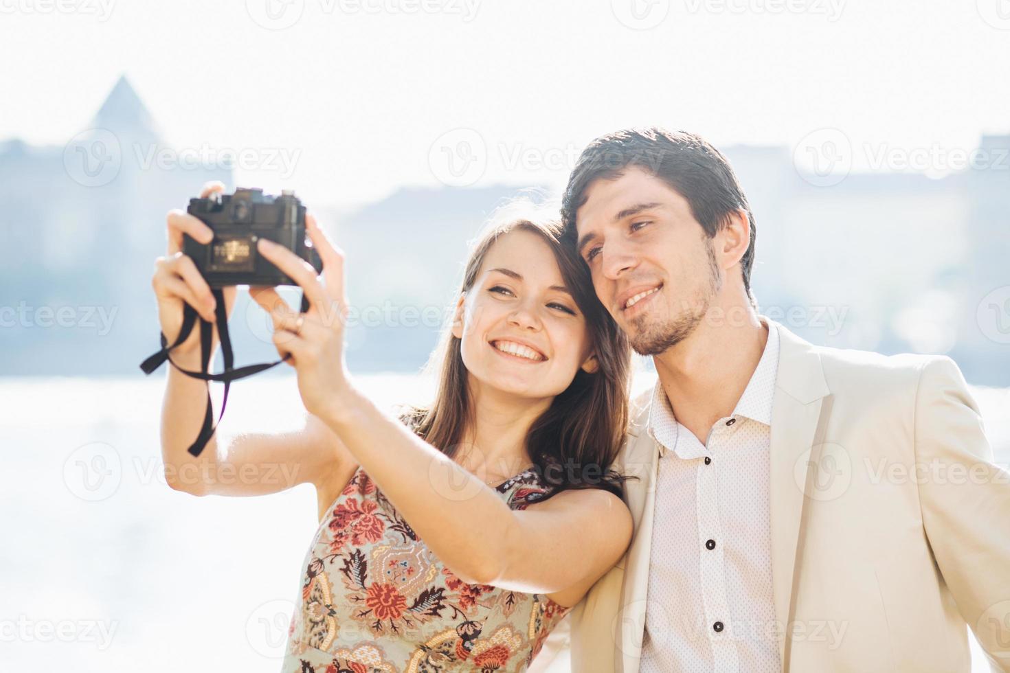 Happy couple in love pose for selfie in modern professional camera, have walk outdoor, explore unknown places in big tourists city, model against wonderful landscape with water in background photo