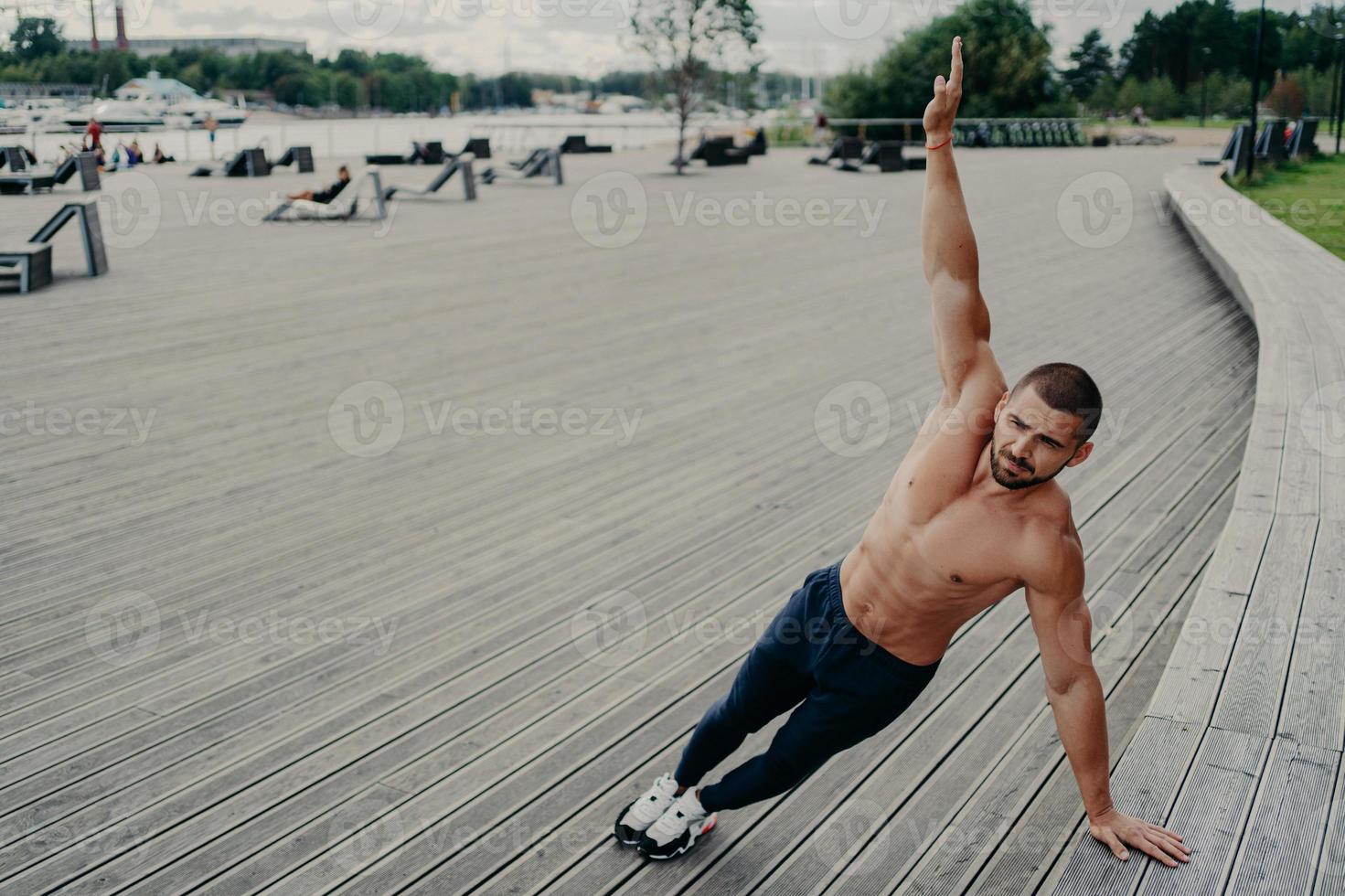 Self determined muscular man dressed in sport clothes stands in side plank, keeps arm raised, has workout outdoor during summer day. Sport, healthy lifestyle, strength and endurance concept. photo