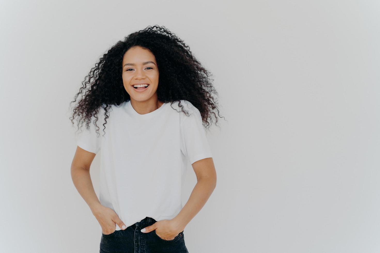 Half length shot of good looking smiling woman laughs at funny joke, has fun, keeps both hands in pockets of jeans, wears white t shirt, has curly fluffy hair, poses indoor, blank space on right side photo