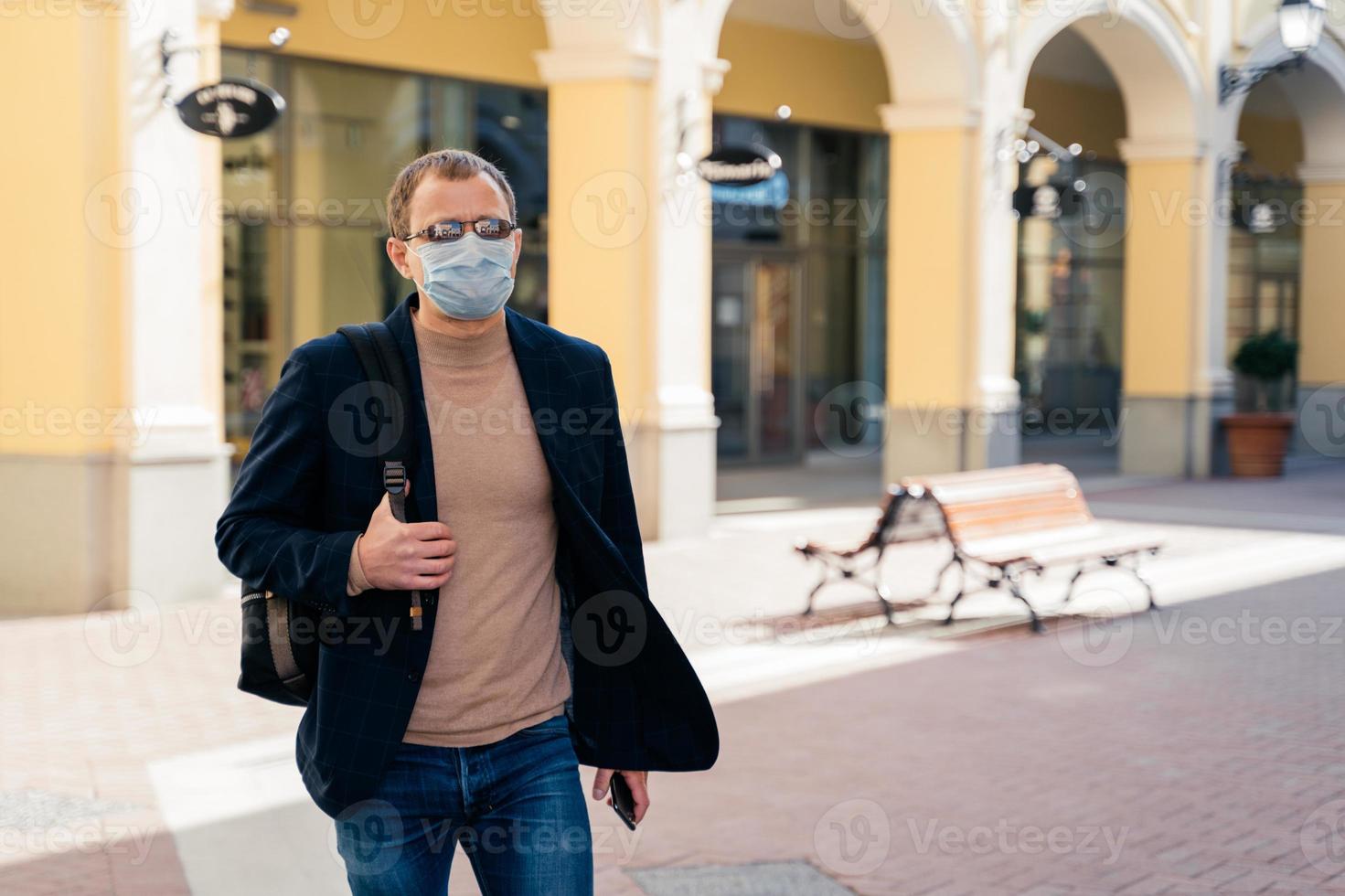 European man wears protective face mask while coronavirus and pandemic, carries rucksack, poses outdoor at bus station, travels during quarantine. Covid-19 virus, epidemic outbreak, public place photo