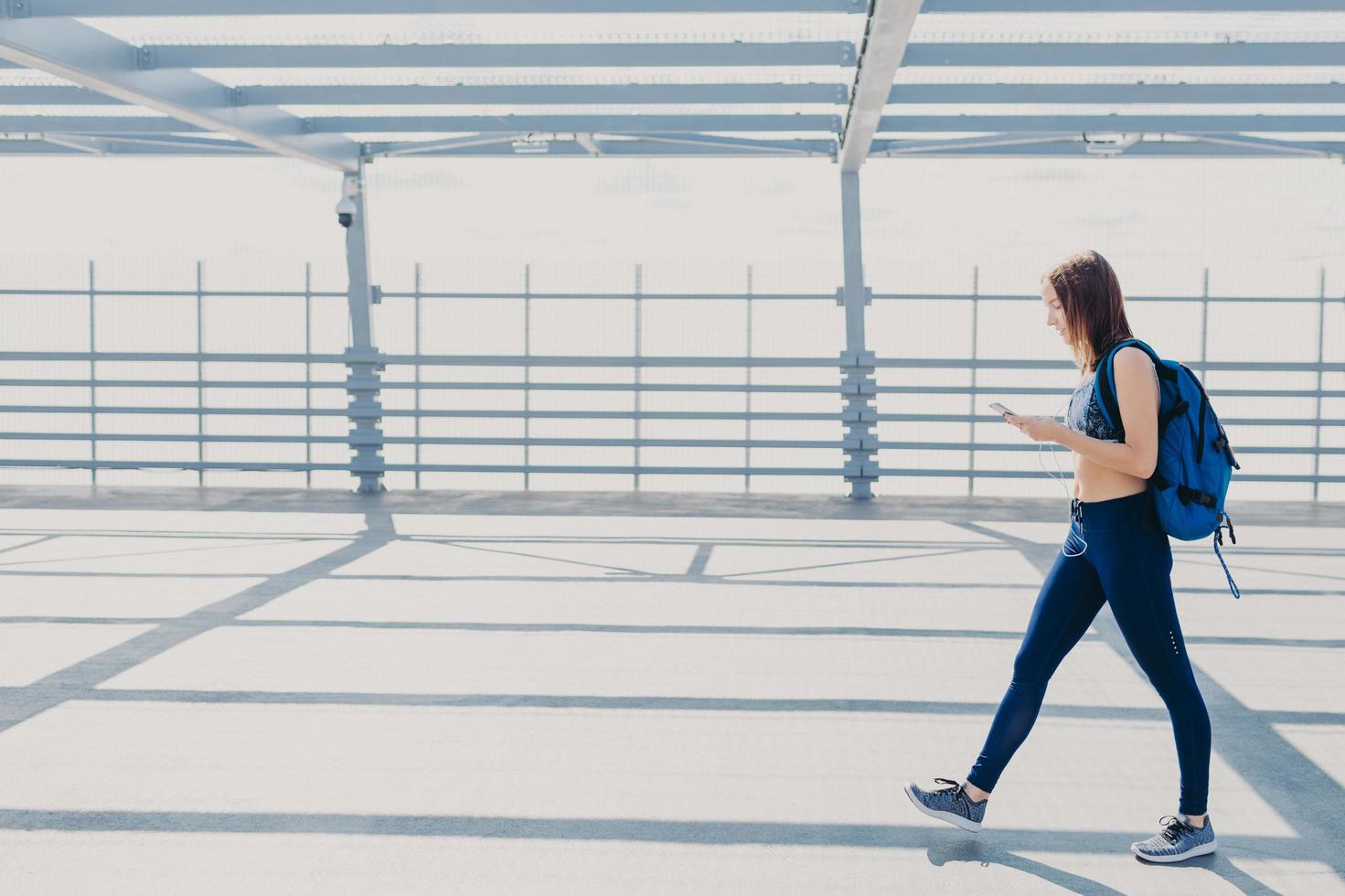 Full length portrait of sporty active female teenager or youngster, wears top, leggings and sneakers, carries rucksack, enjoys pleasant music in playlist, uses modern cell phone and earphones photo