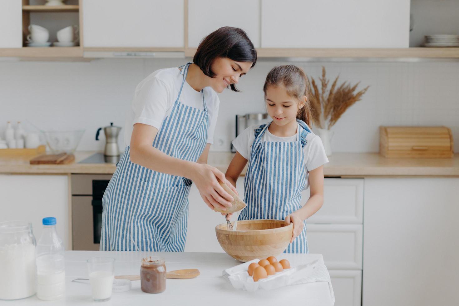 Daughter and mother love cooking, dressed in striped aprons, prepare delicious dinner, use different ingredients, have kitchen routine, stand at home. Children, motherhood, culinary concept. photo