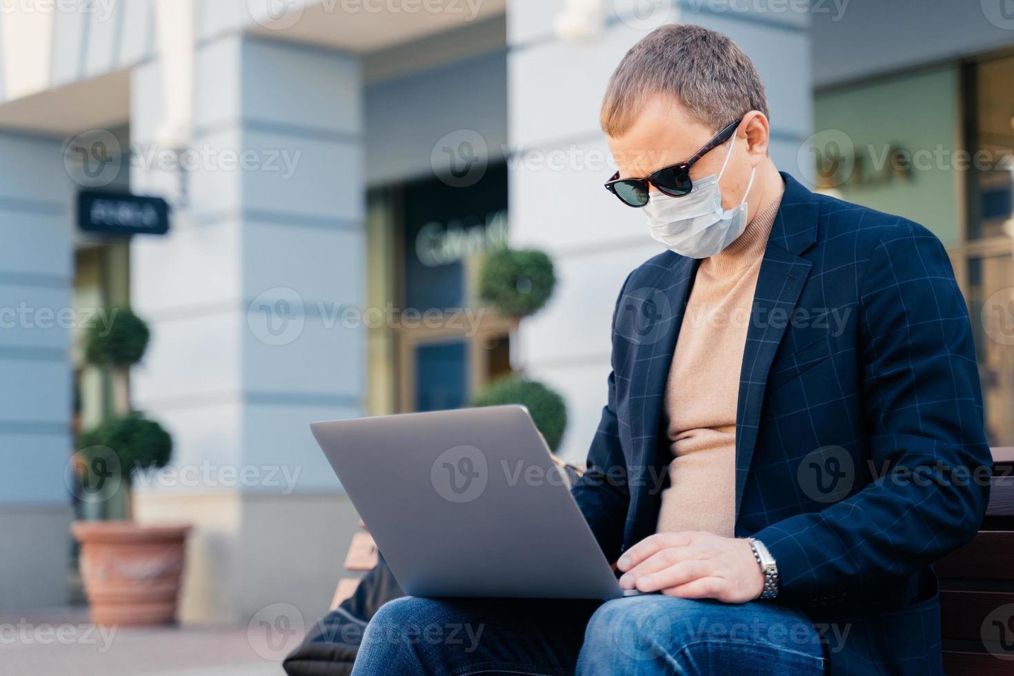 Concentrated businessman poses outdoor near bus station, uses laptop computer for distance work, wears protective mask to avoid coronavirus. People, technology, travel and virus outbreak concept photo