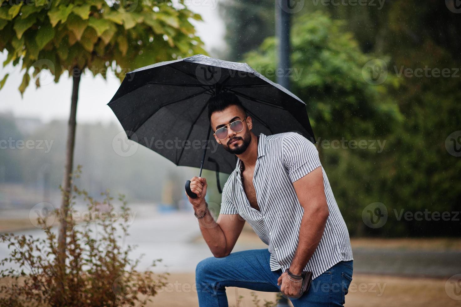 Fashionable tall arab beard man wear on shirt, jeans and sunglasses with umbrella posed at rain on park square. photo
