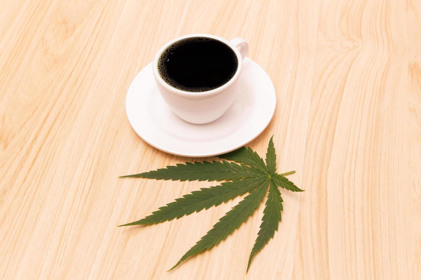 Hot black coffee in a cup with a white saucer and coffee beans sprinkled on a wooden table and decorated with hemp leaves, cannabis coffee relaxing drink concept. photo