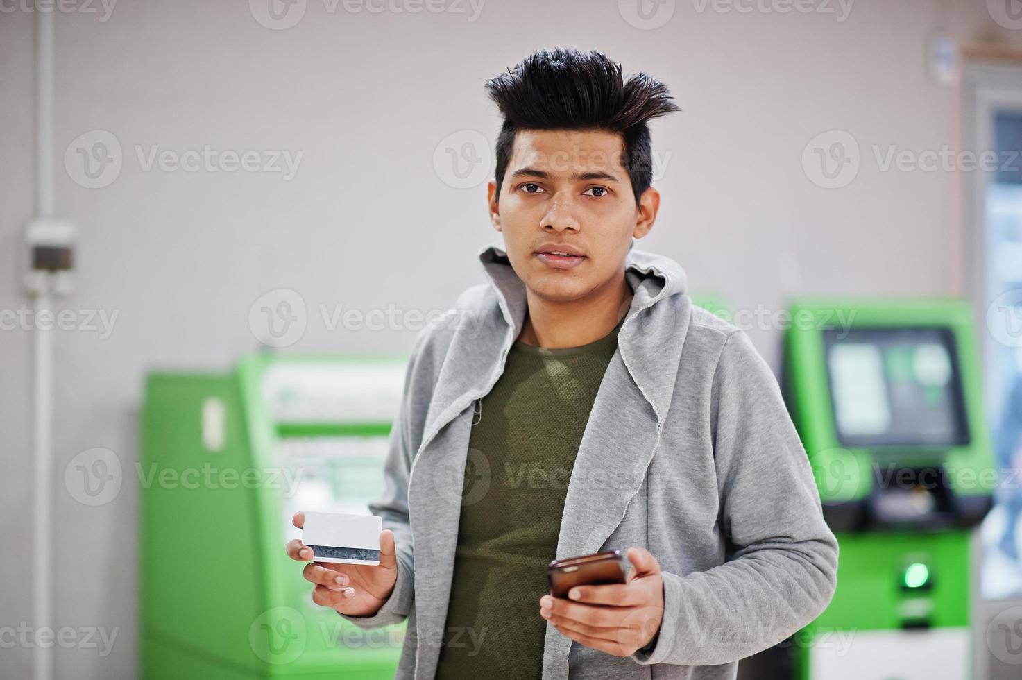 Young stylish asian man with mobile phone and credit card in hands against row of green ATM. photo