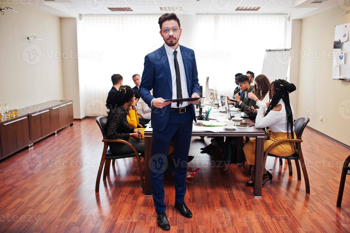 Face of handsome arabian business man, holding clipboard on the background of business peoples multiracial team meeting, sitting in office table. photo