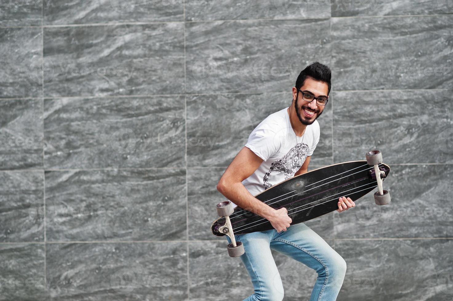 Street style arab man in eyeglasses with longboard posed against gray wall, like he play guitar. photo