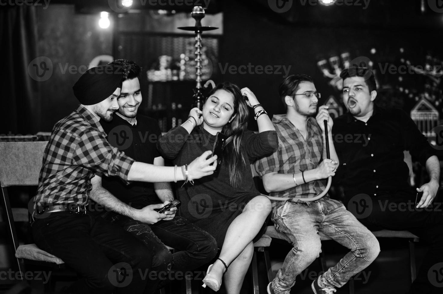 Group of indian friends sitting at lounge bar, having fun and rest, smoking hookah and looking on mobile phones. photo