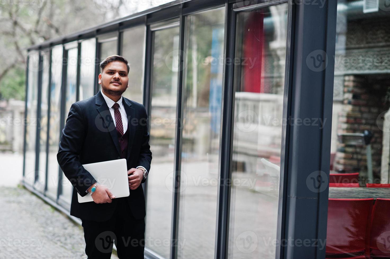 hombre de negocios indio con estilo en ropa formal con portátil en las manos de pie contra las ventanas en el centro de negocios. foto