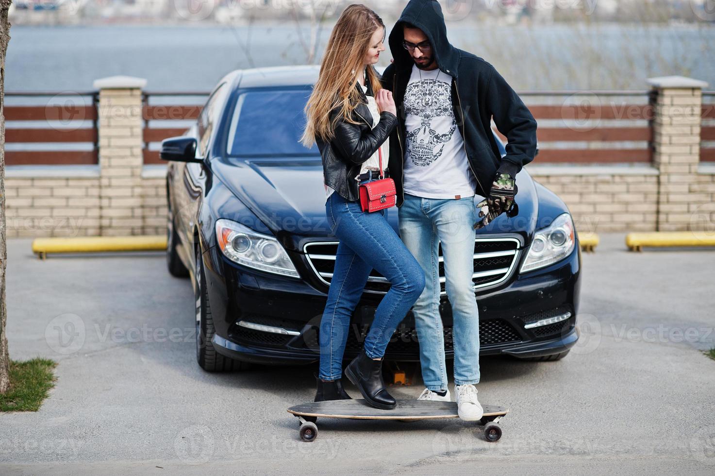 Cool multiracial couple hugs together with longboard against black elegant sport car. photo