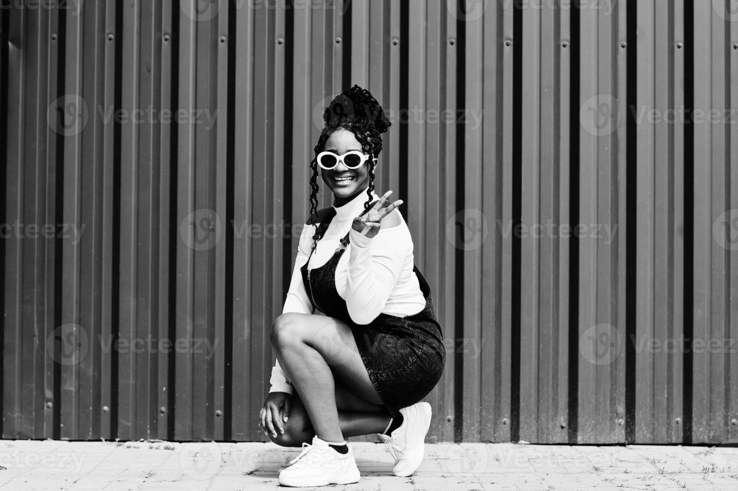 African woman in denim overalls skirt, white sunglasses crouching and show two fingers against green steel wall. photo