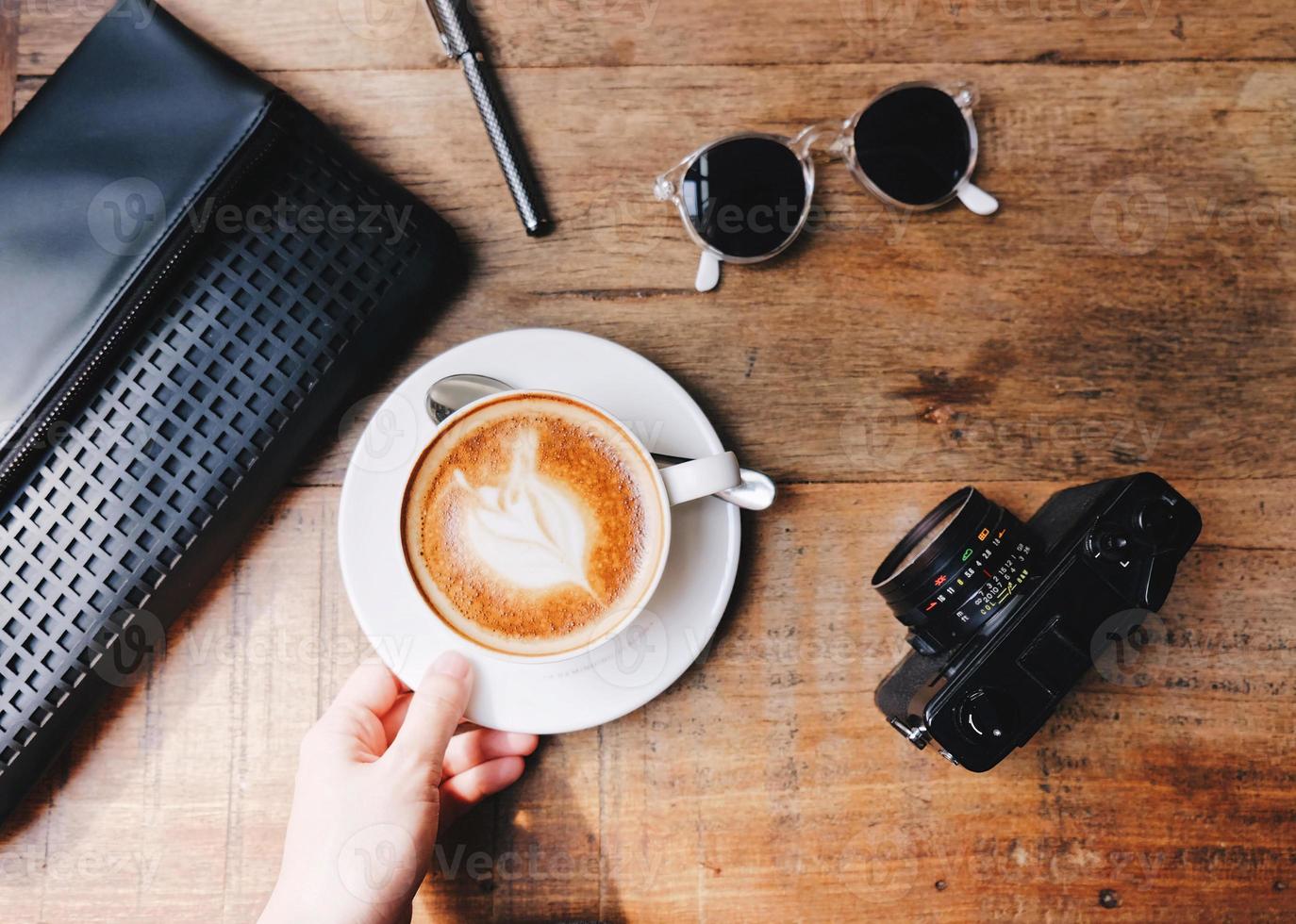 Top view of hand holding hot coffee on wooden table with woman bag with camera and sunglasses photo