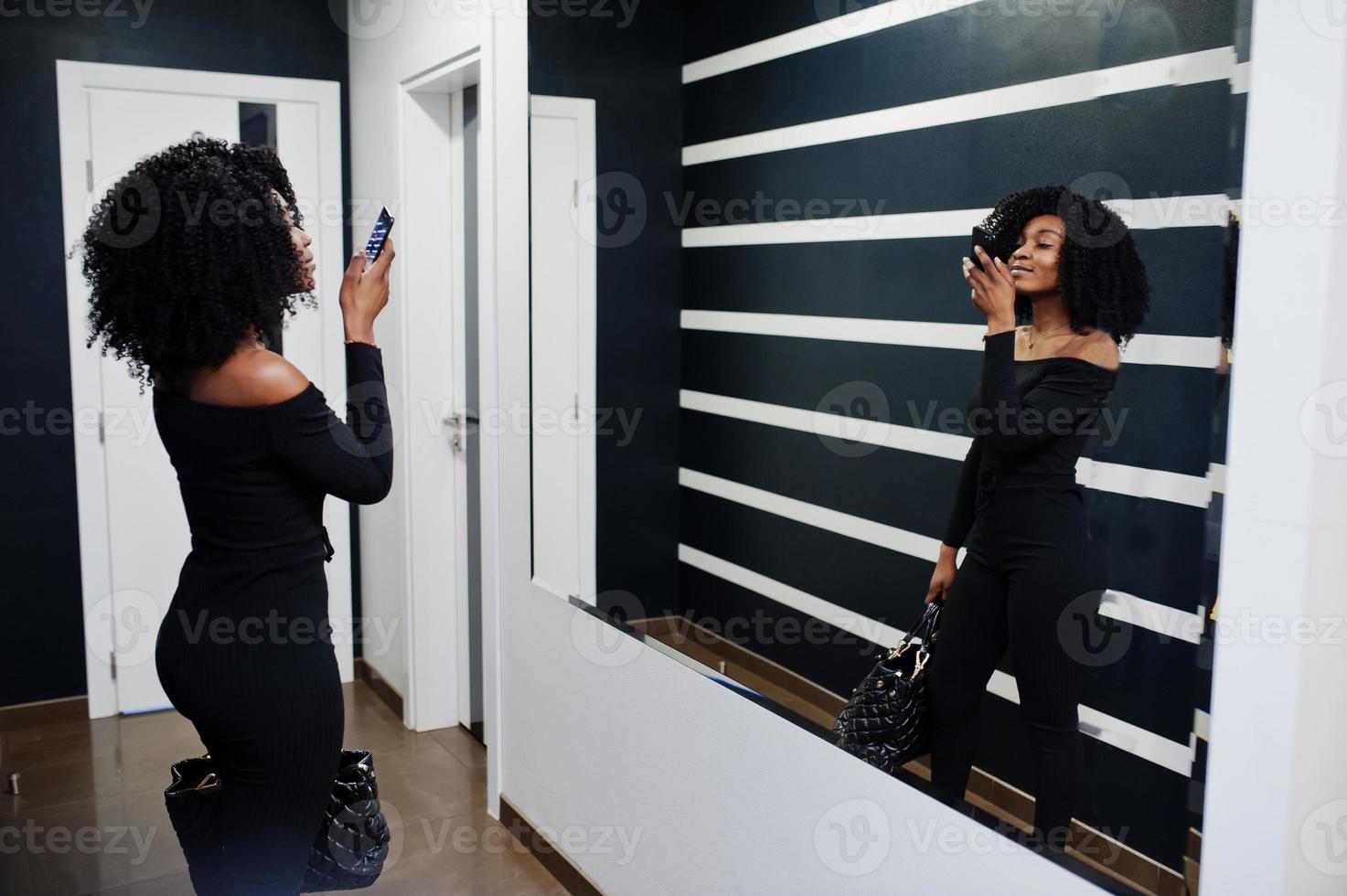 Fashionable young beautiful african american woman with afro hairstyle and hangbag posing wear in elegant black. Making photo at mirror by phone.