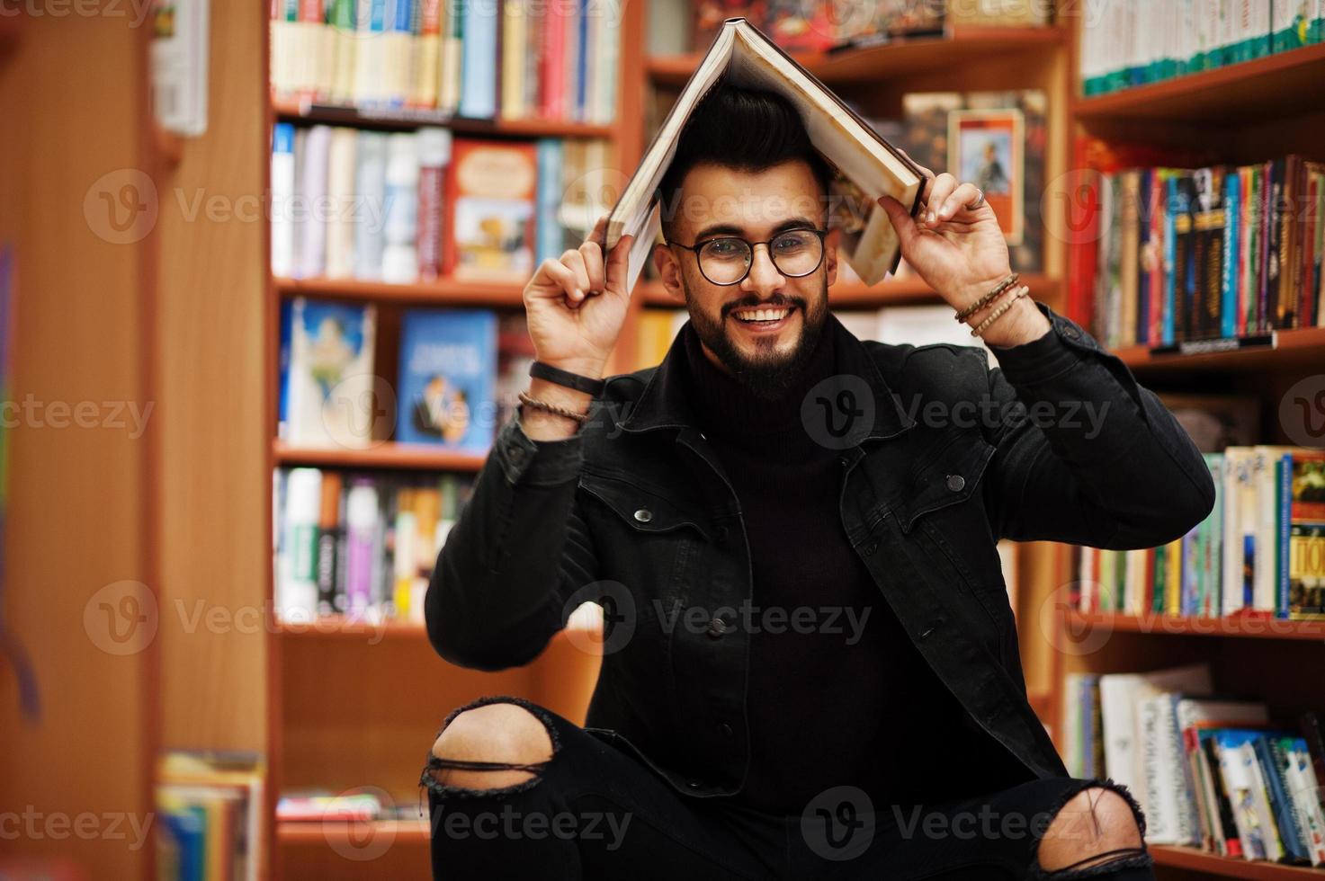 estudiante árabe alto e inteligente, vestido con chaqueta negra de jeans y anteojos, sentado en la biblioteca y cubriendo la cabeza con un libro. foto