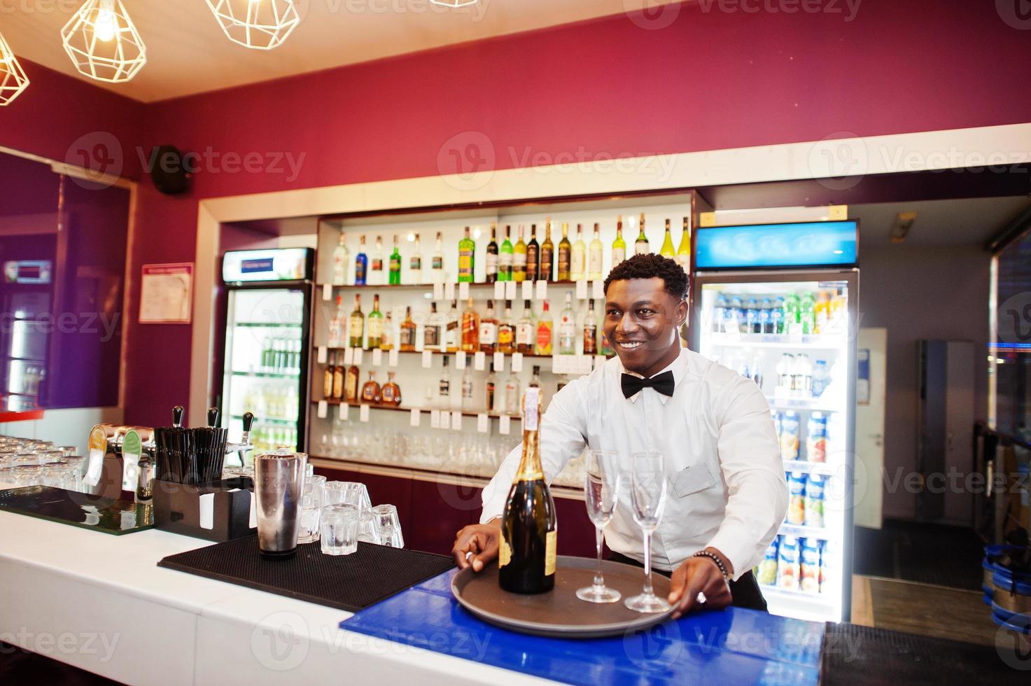 African american bartender at bar holding champagne with glasses on tray. photo