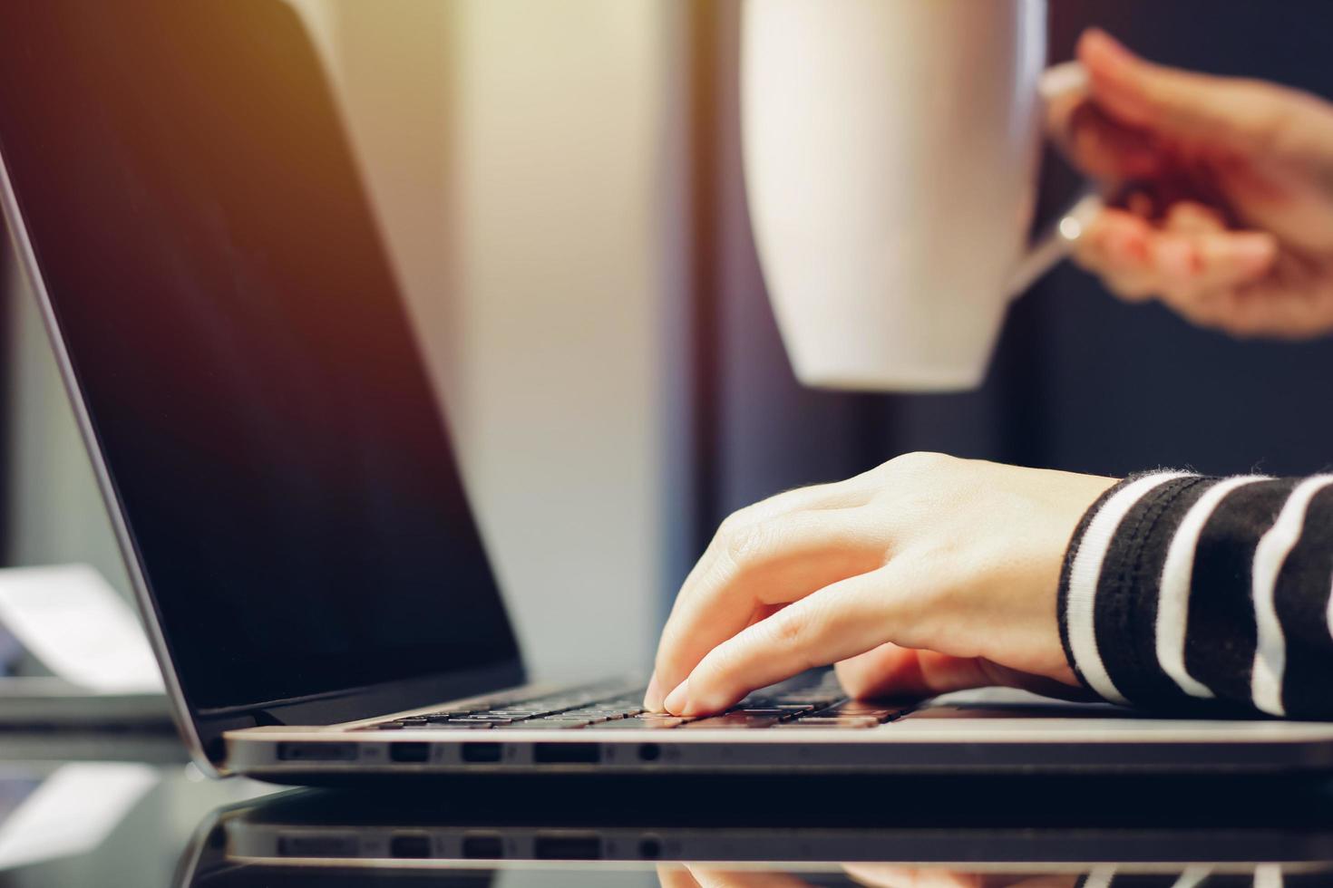 manos femeninas escribiendo en el teclado de la computadora portátil mientras sostiene una taza de café, trabajando en el concepto de casa foto