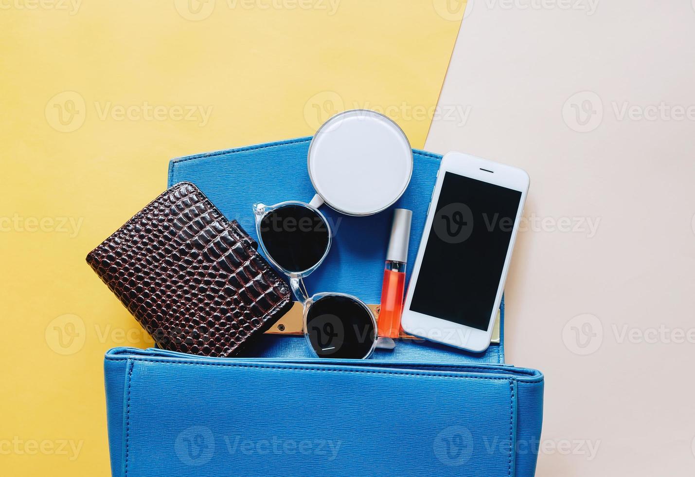 Flat lay of blue leather woman bag open out with cosmetics, accessories, wallet and smartphone on yellow background with copy space photo