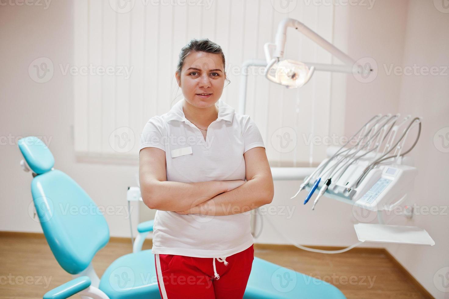 retrato de mujer dentista cruzó los brazos de pie en su oficina de odontología cerca de la silla. foto