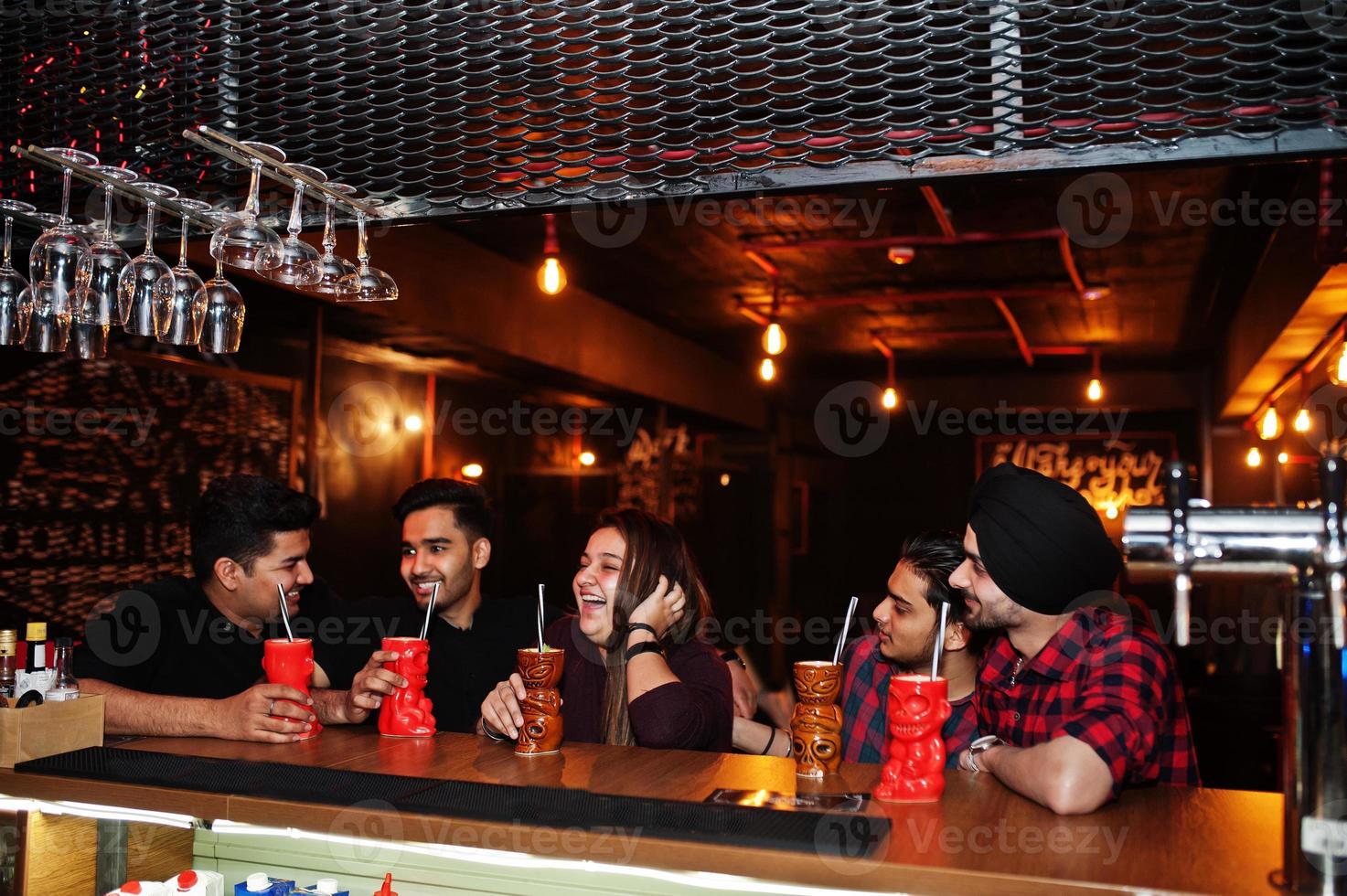 Group of indian friends having fun and rest at night club, drinking cocktails near bar counter. photo