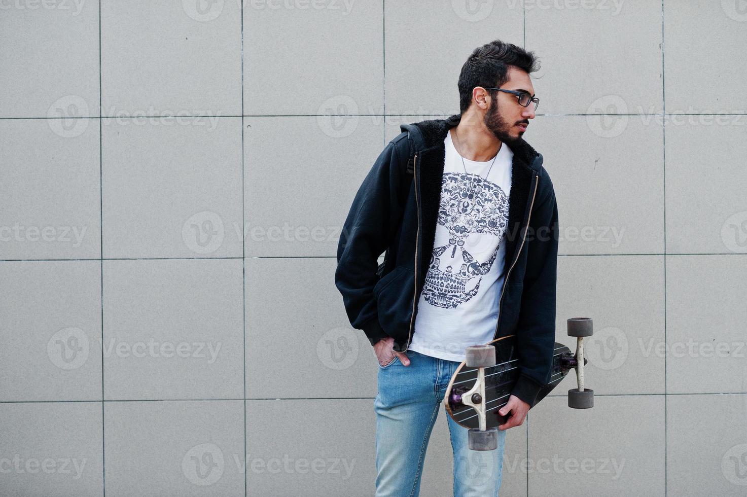 Street style arab man in eyeglasses with longboard posed against gray wall. photo