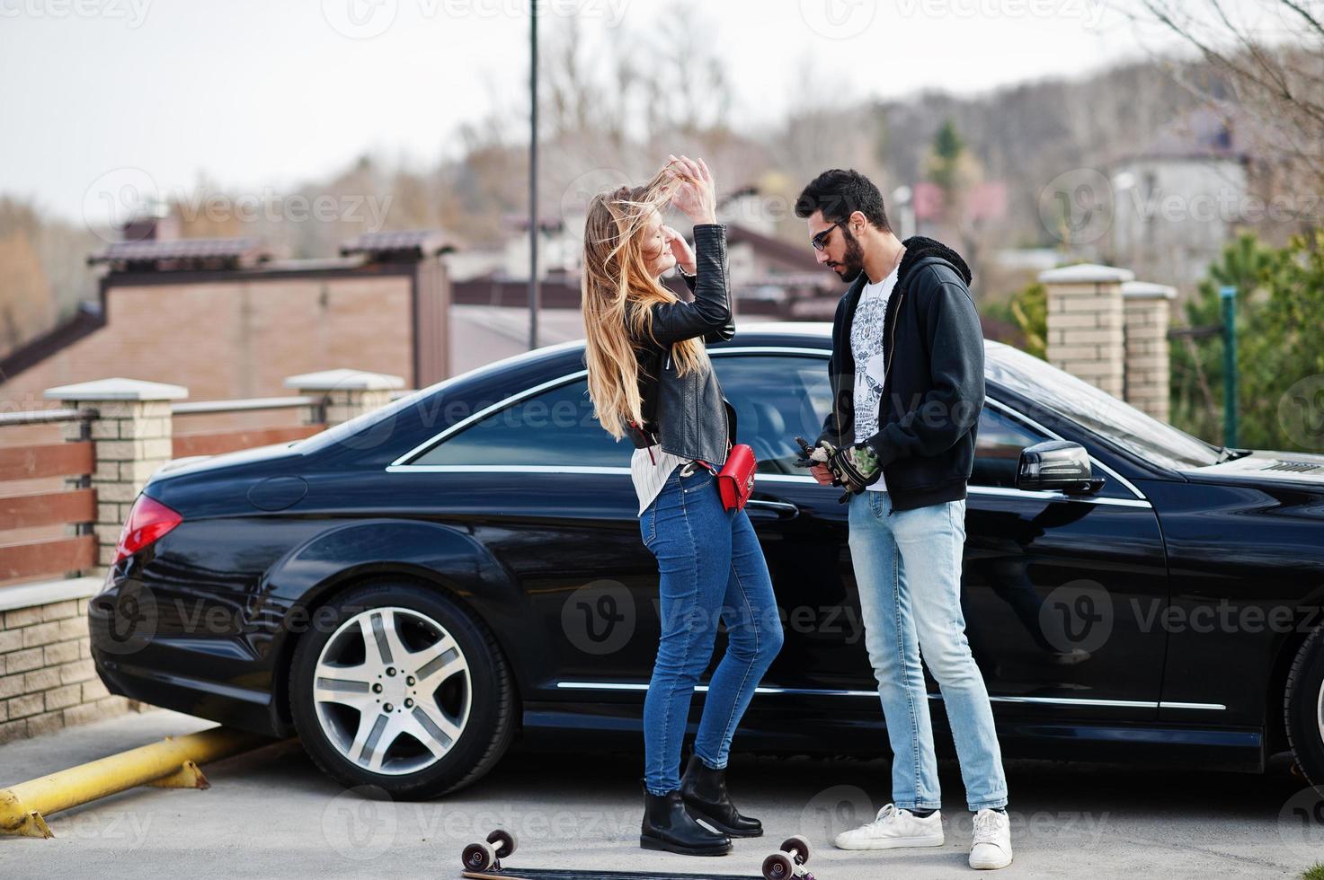 Cool multiracial couple hugs together with longboard against black elegant sport car. photo