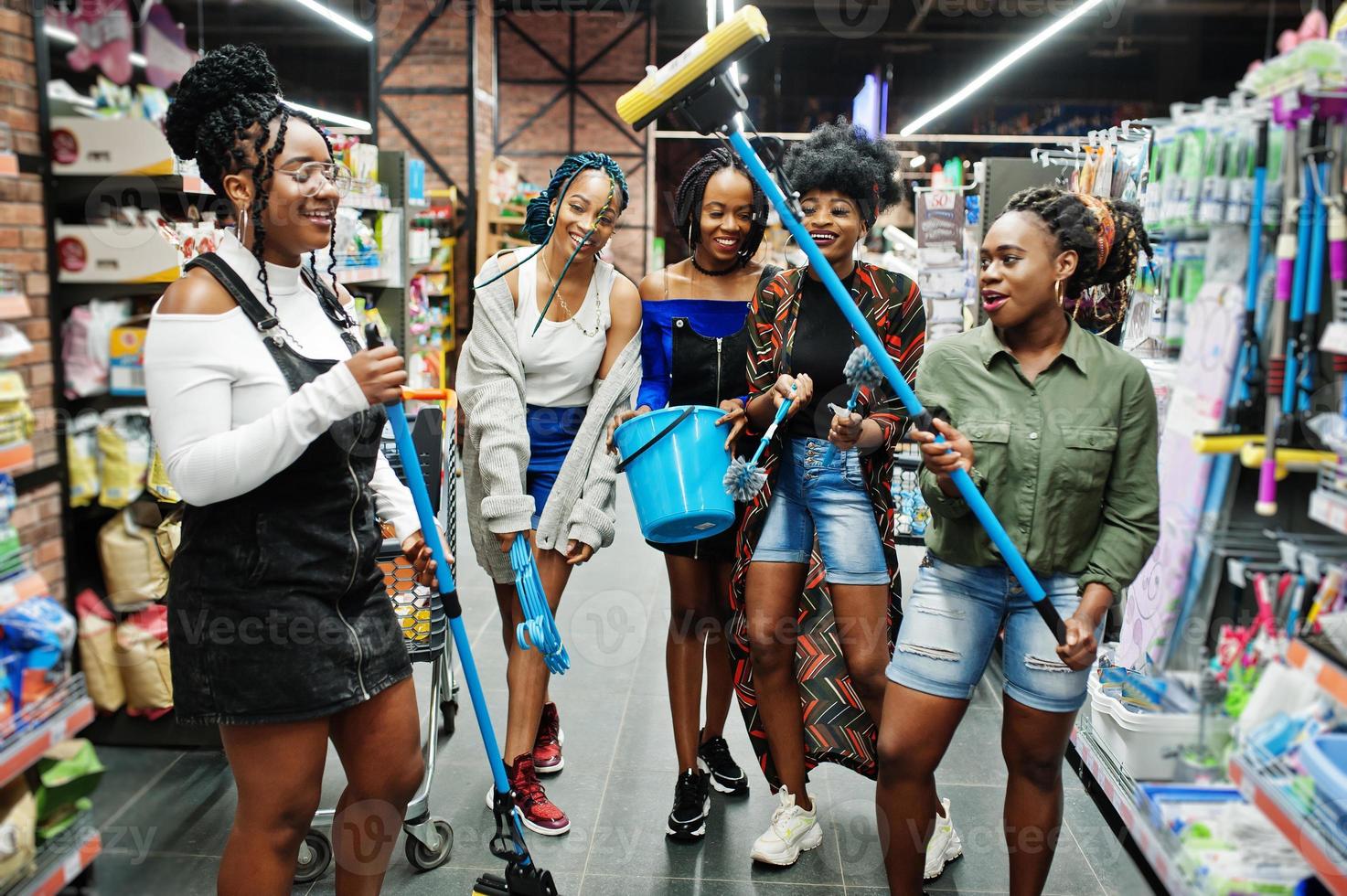 Group of five african womans with dust mop, toilet brush and bucket having fun in household cleaning items department in supermarket. photo