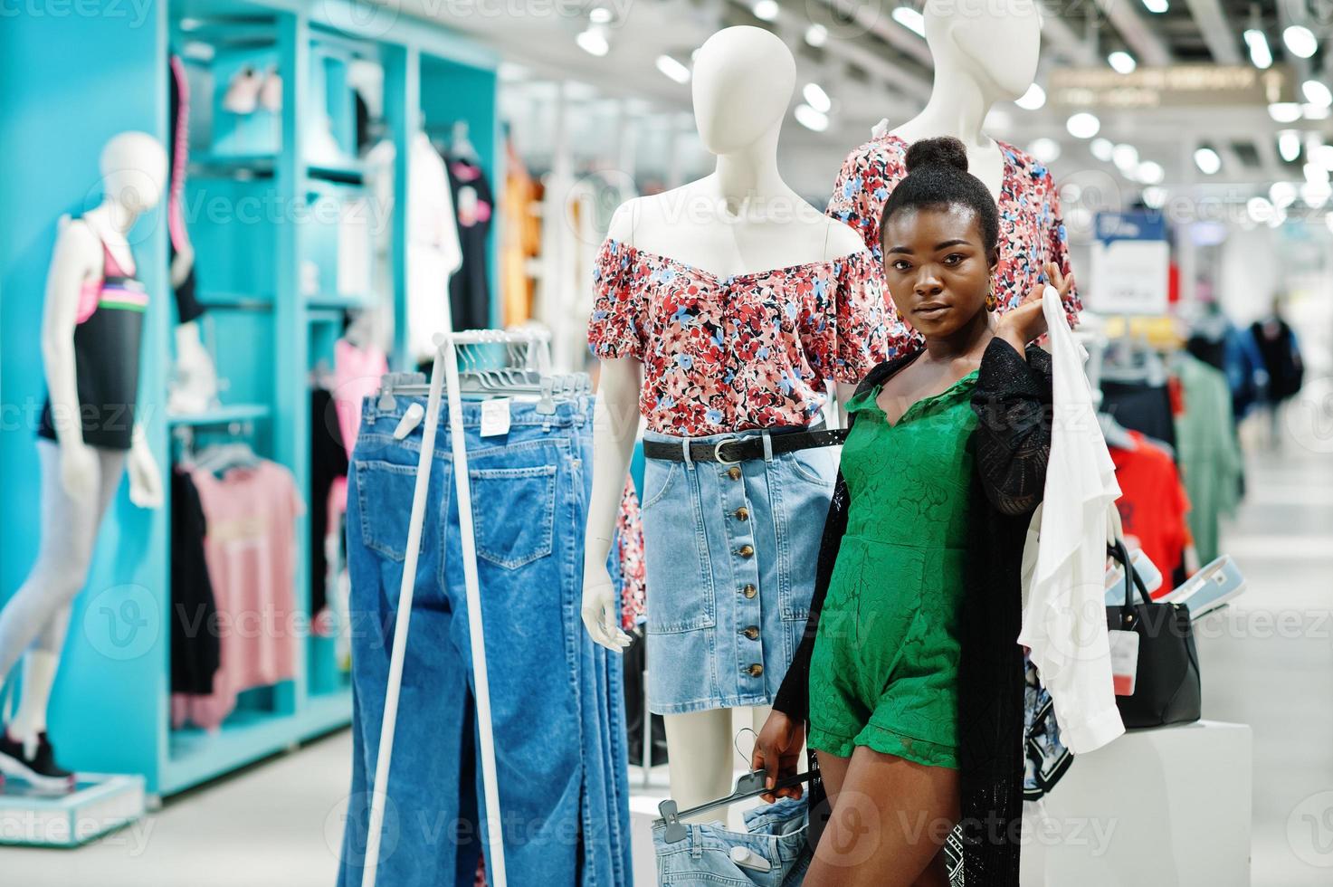 Young fashionable and sexy afro woman in green combidress shopping at clothes store. She hold shirt and shorts on hangers against mannequin. photo