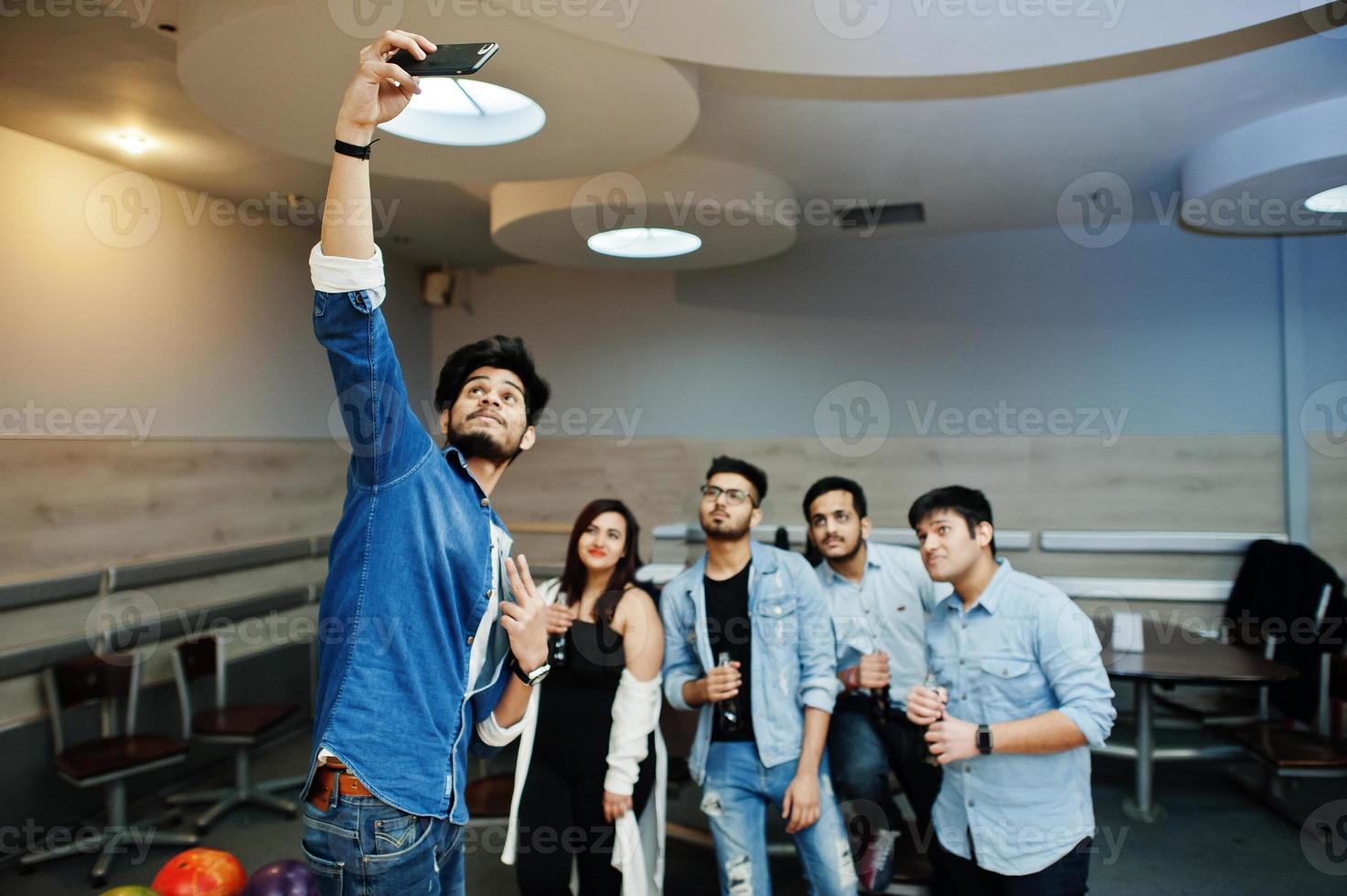 Group of five south asian peoples having rest and fun at bowling club. Making selfie by phone and holding cold soda drinks from glass bottles. photo