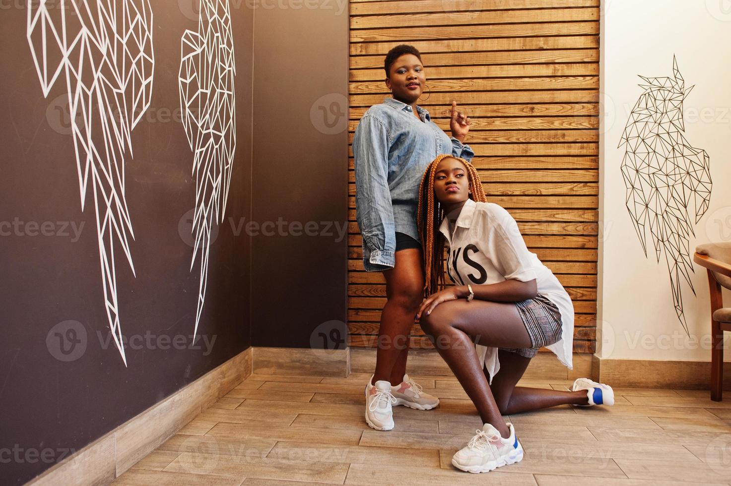 Two african womans in stylish casual clothes posing against wooden wall sat down and dance twerk. photo
