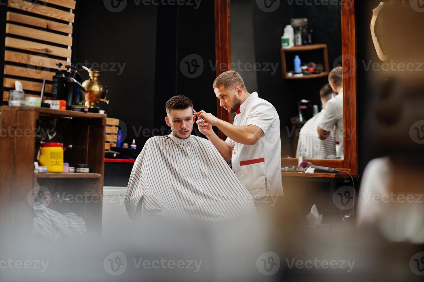 Young bearded man getting haircut by hairdresser while sitting in chair at barbershop. Barber soul. photo