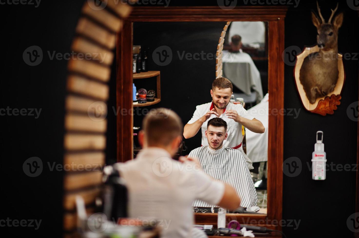Young bearded man getting haircut by hairdresser while sitting in chair at barbershop. Barber soul. photo
