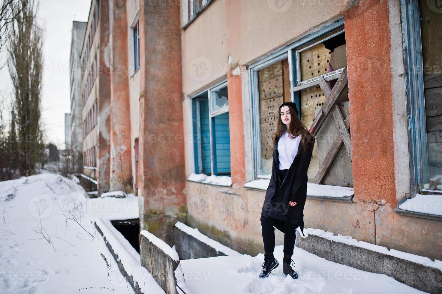 modelo morena de piernas largas de moda en un largo manto negro posado al aire libre en el día de invierno contra la vieja pared de grunge con ventanas rotas. foto