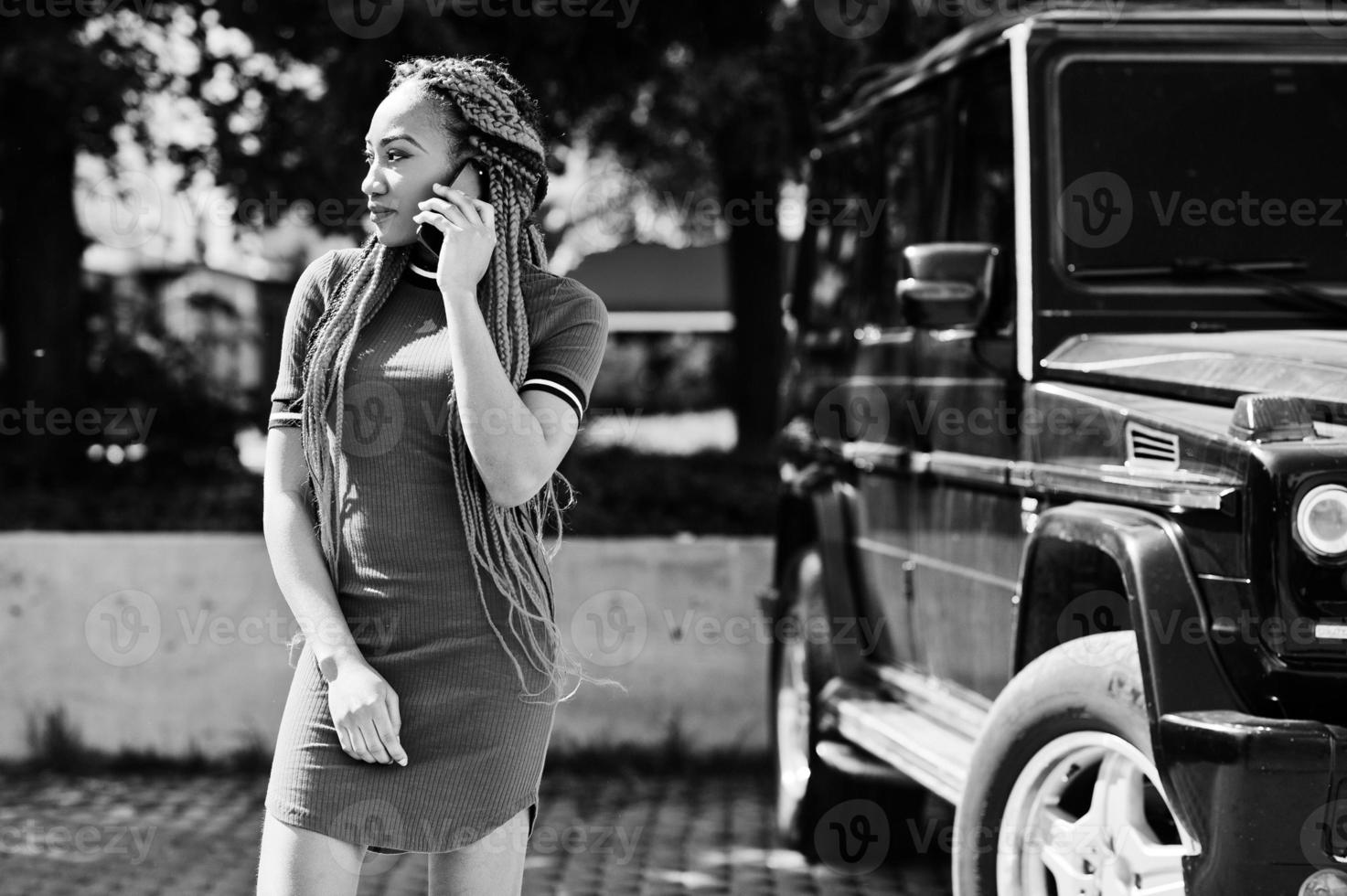 Cute and slim african american girl in red dress with dreadlocks background black mafia suv car and speking on mobile phone. Stylish black model. photo