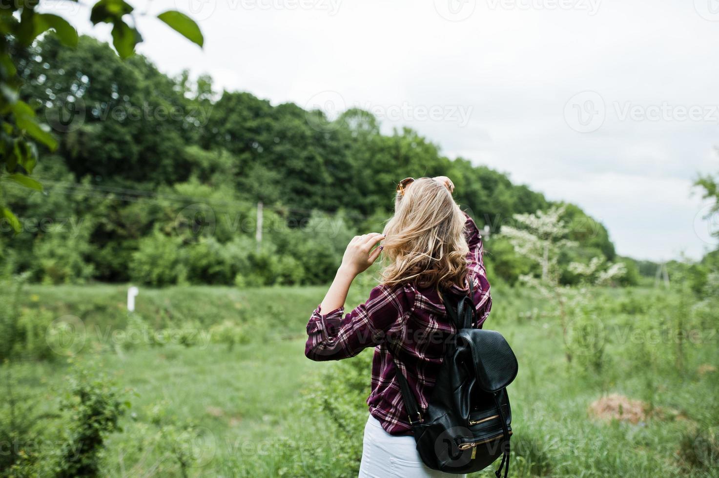 retrato de una atractiva chica rubia con camisa de tartán con mochila caminando por el campo. foto