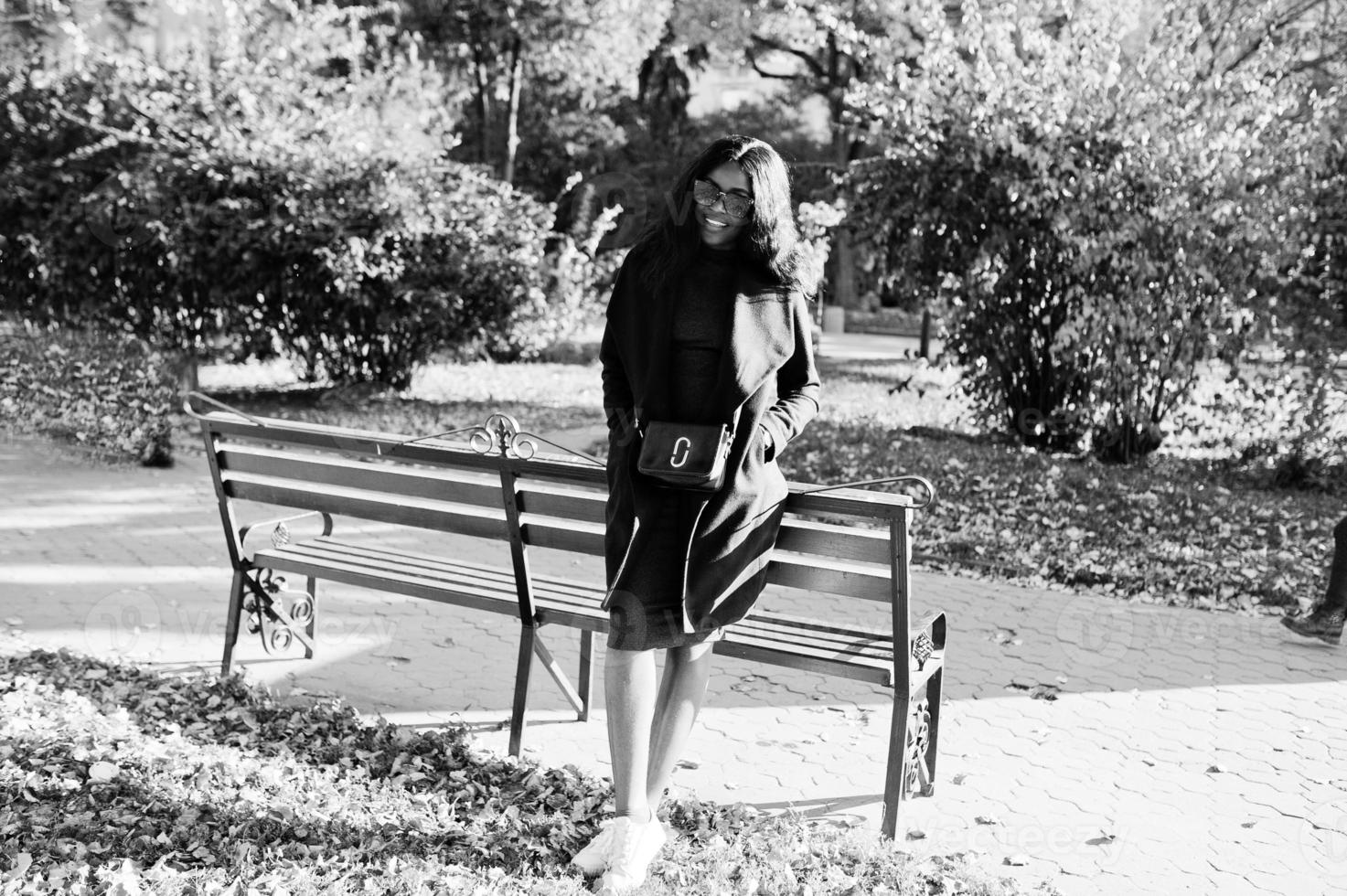 Stylish african american girl in blue coat and sunglasses posed at sunny autumn day at bench. Africa model woman. photo