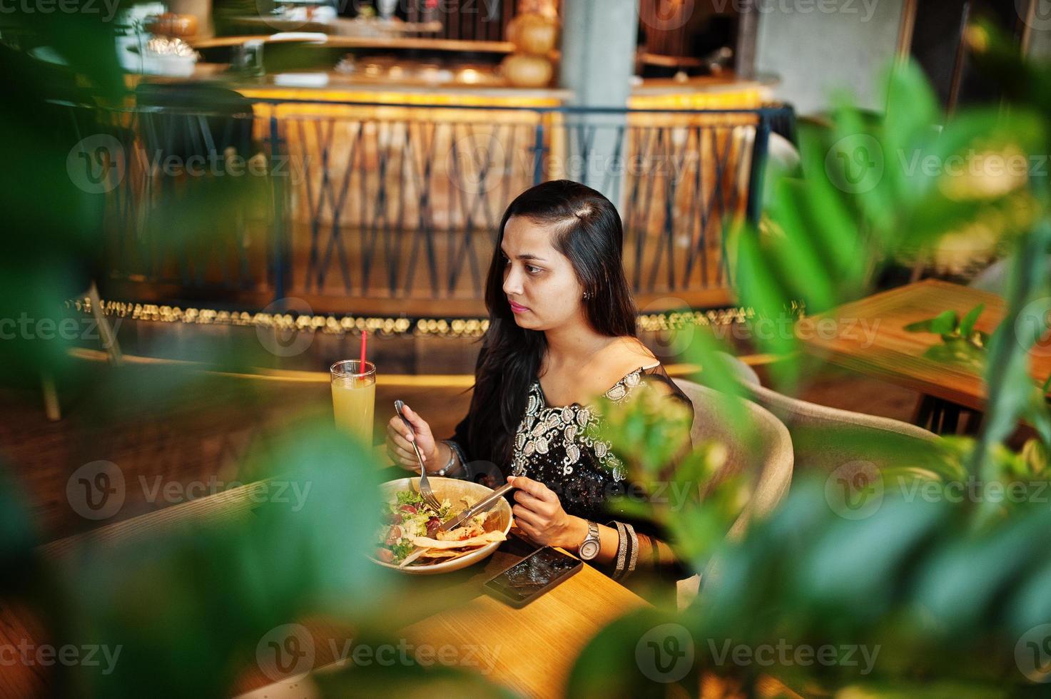 Pretty indian girl in black saree dress posed at restaurant, sitting at table with juice and salad. photo