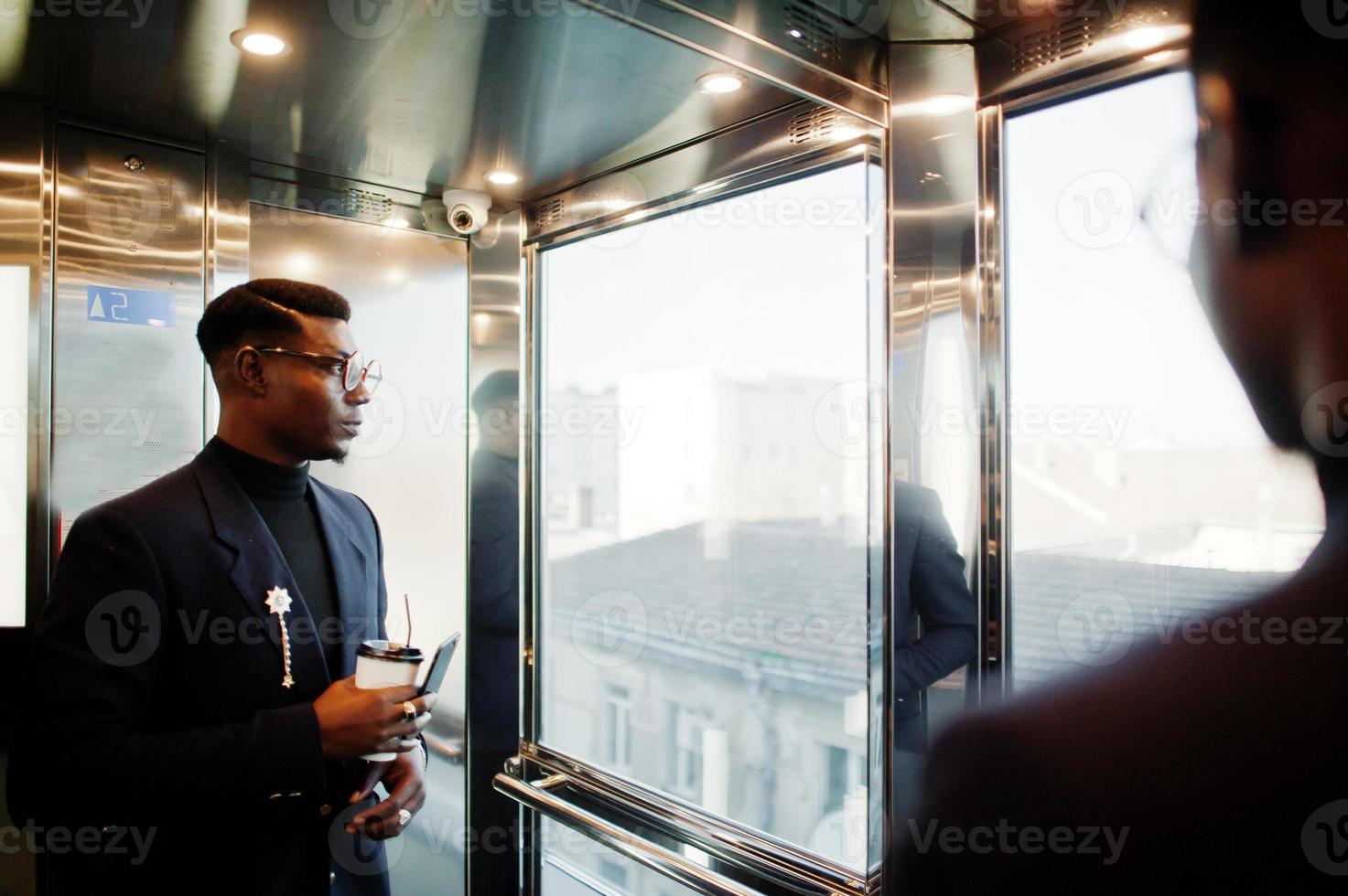 Fashionable african american man in suit and glasses with mobile phone and cup of coffee at hands posed inside elevator. photo