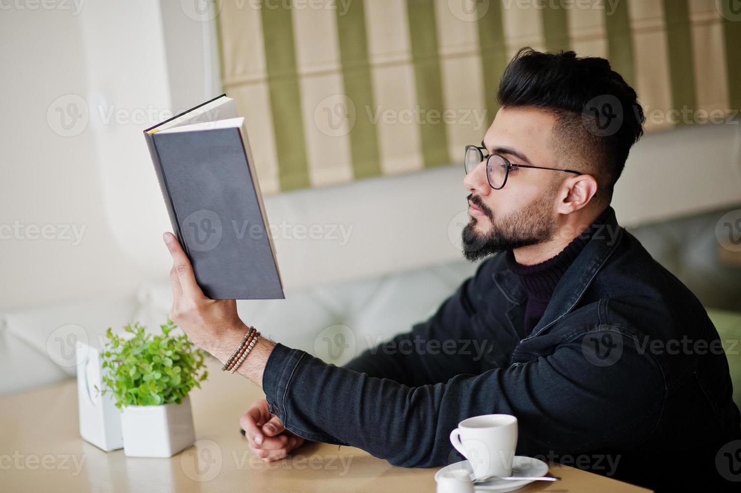 Arab man wear on black jeans jacket and eyeglasses sitting in cafe, read book and drink coffee. Stylish and fashionable arabian model guy. photo