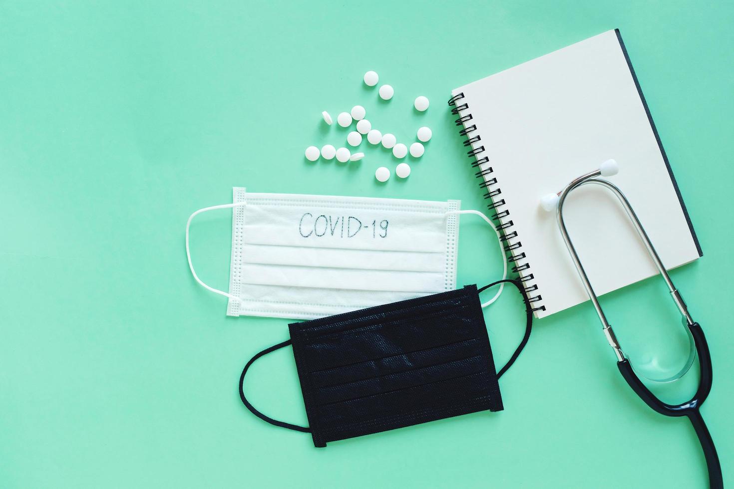 Flat lay of medicine pills, mask, blanl notebook and stethoscope on green background, health care concept and prevent the spread of pandemic Covid-19 and Coronavirus photo