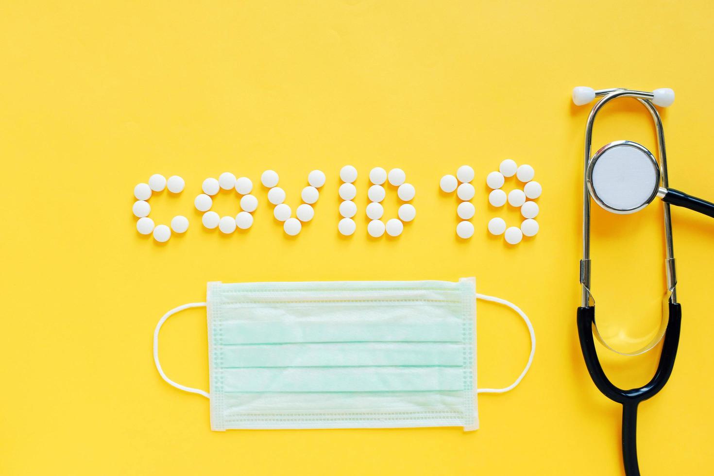 Flat lay of medicine pills shape as word covid 19 with mask and stethoscope on yellow background, health care concept and prevent the spread of pandemic Covid-19 and Coronavirus photo