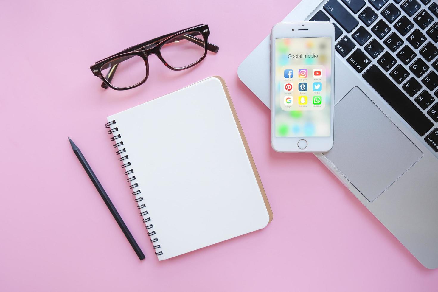 Group of Popular Social networks icons showing on Apple iPhone 6s screen with laptop and blank notebook on pink background, Social media are most popular tool for communication. photo