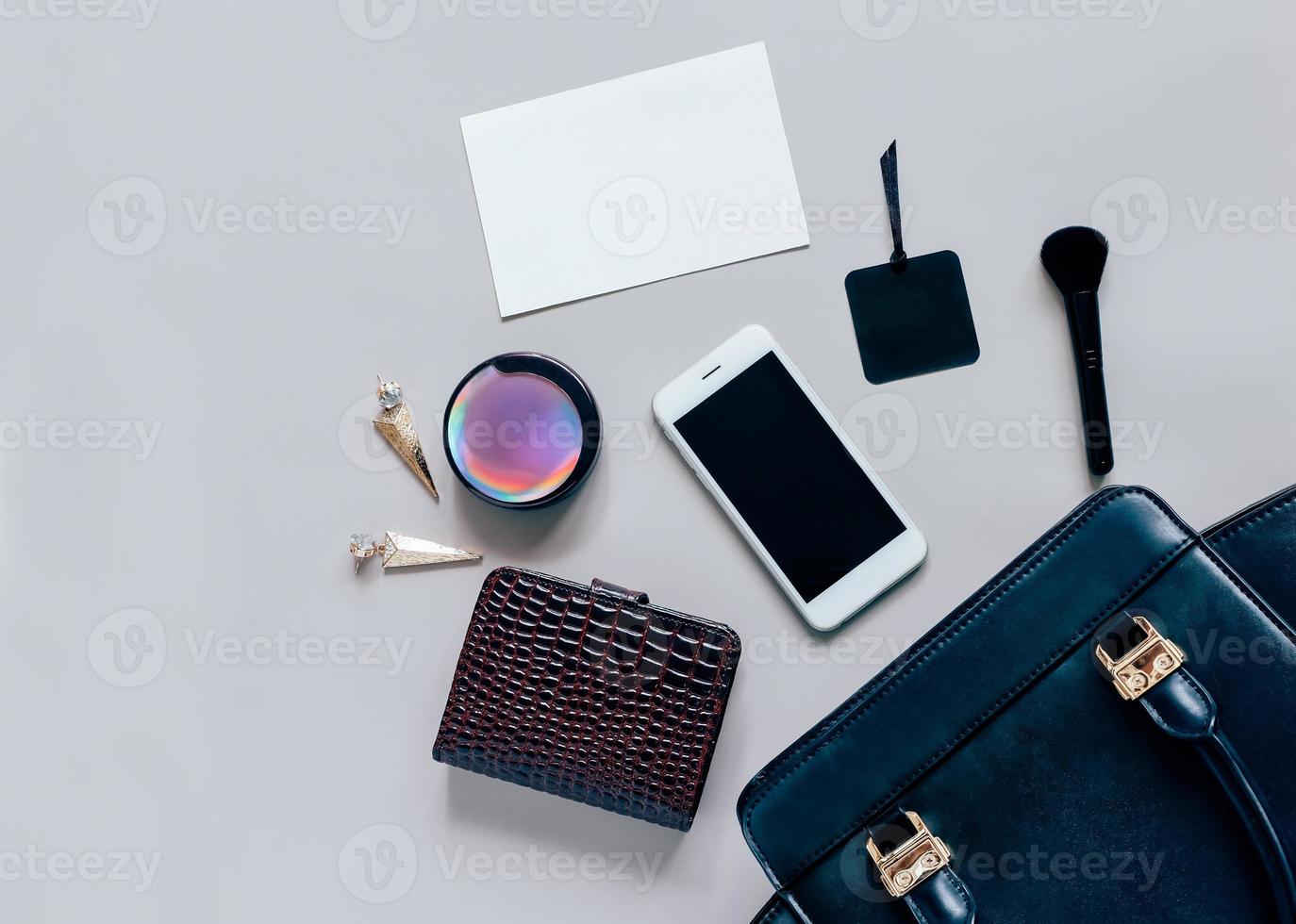 Flat lay of black leather woman bag open out with cosmetics, accessories and smartphone and blank card of paper on grey background with copy space photo