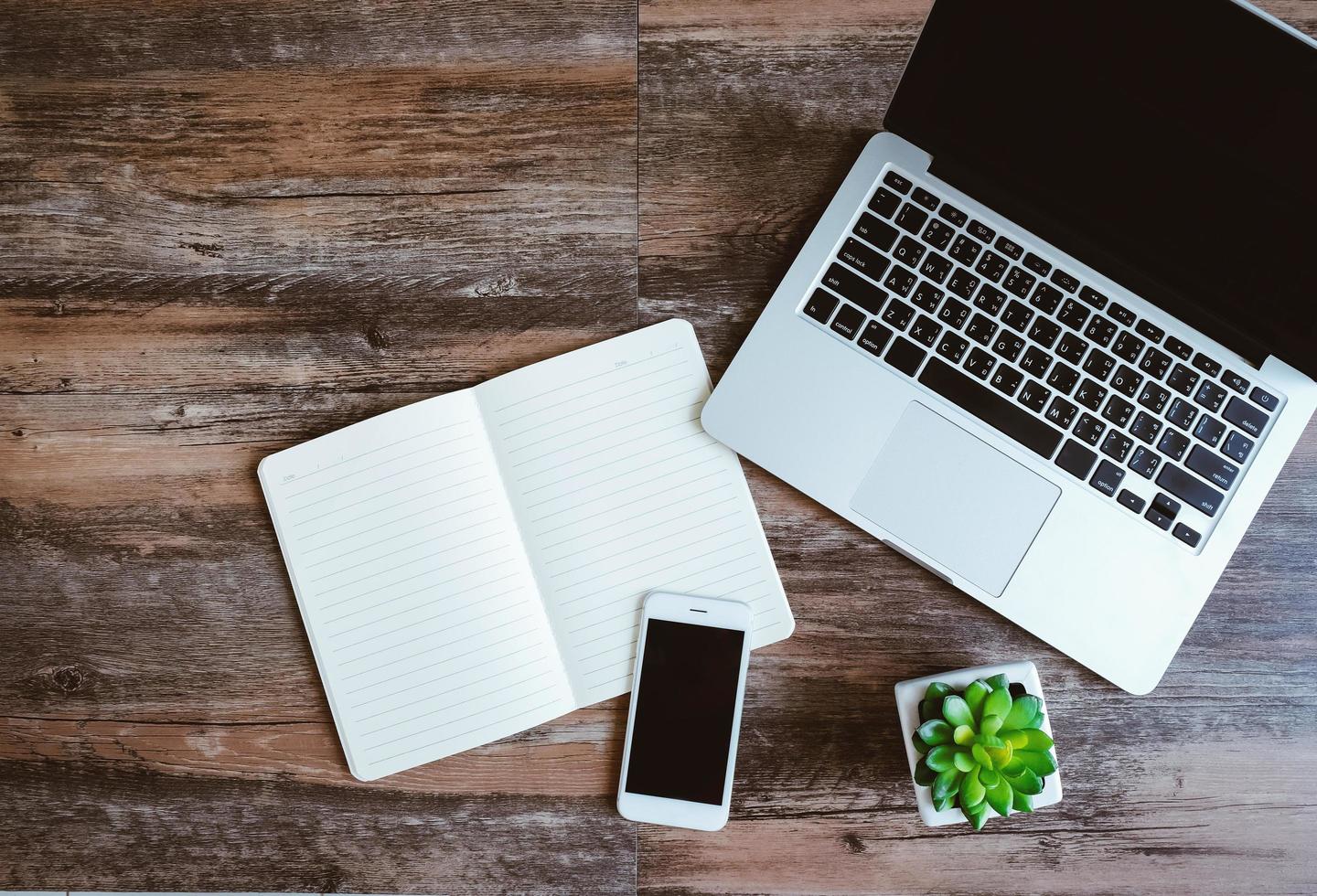 Flat lay photo of workspace desk with laptop, smartphone, blank notebook and green plant with copy space wooden background