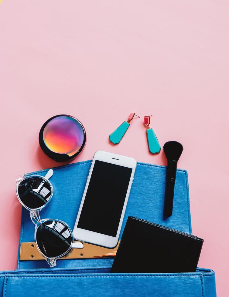 Flat lay of blue leather woman bag open out with cosmetics, accessories, wallet and smartphone on pink background with copy space photo