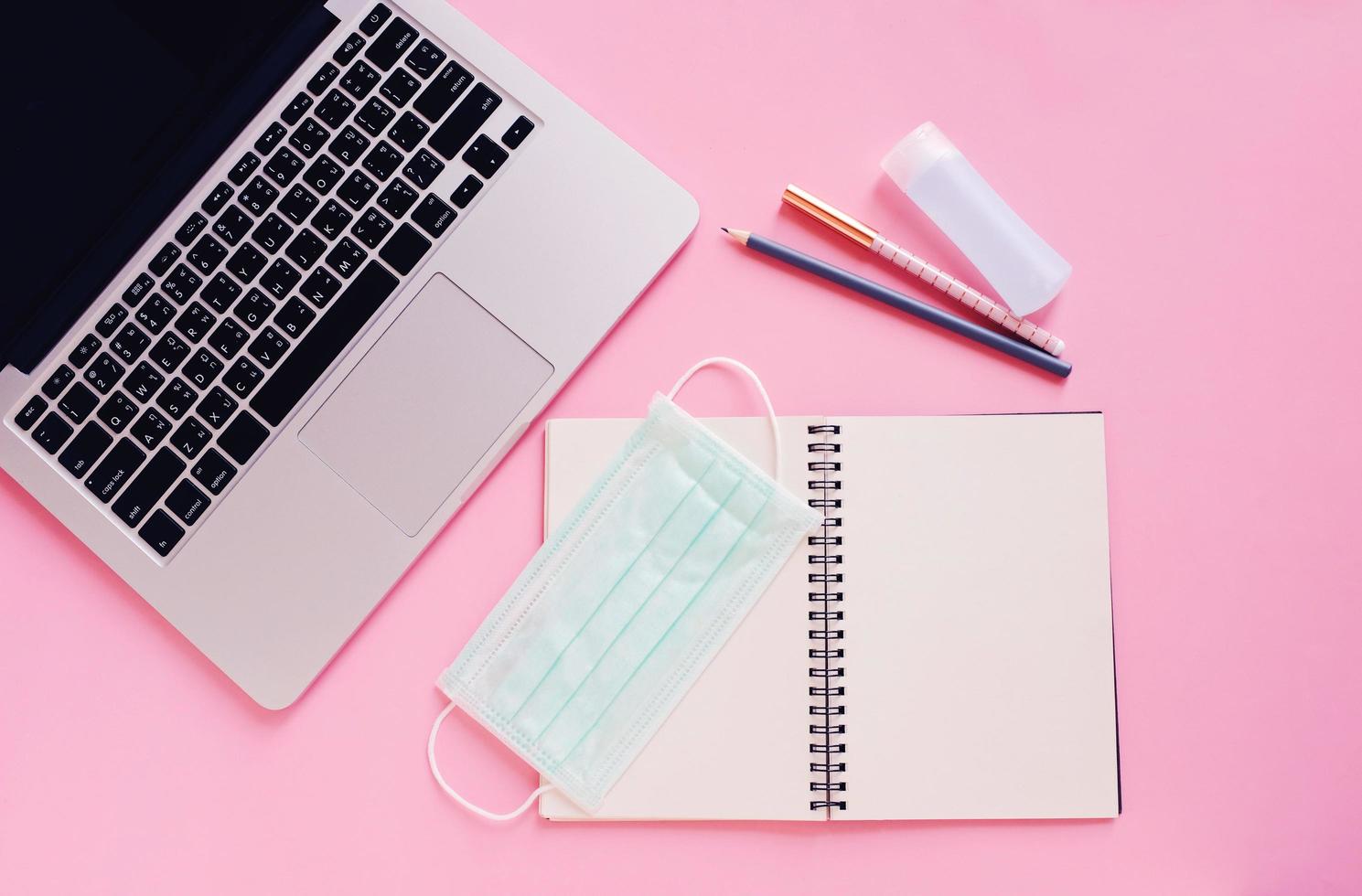 Top view flat lay of workspace desk with laptop, blank notebook, medical masks and alcohol gel on bright pink background, work from home and self quarantine concept photo