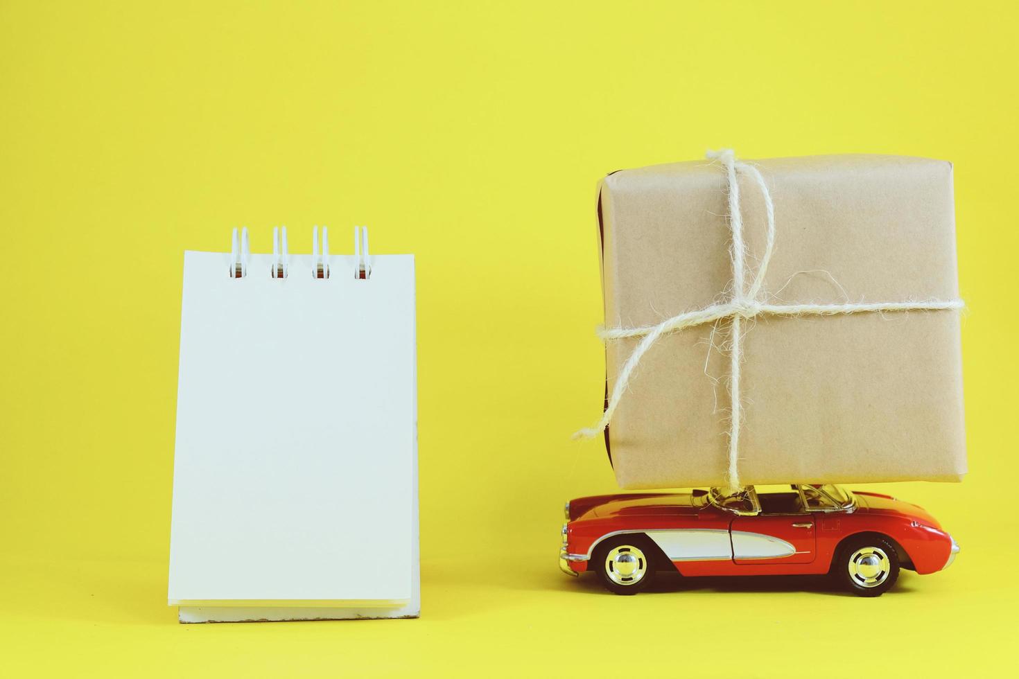 Red retro toy car delivering gift box for holiday season on yellow background with blank notebook photo