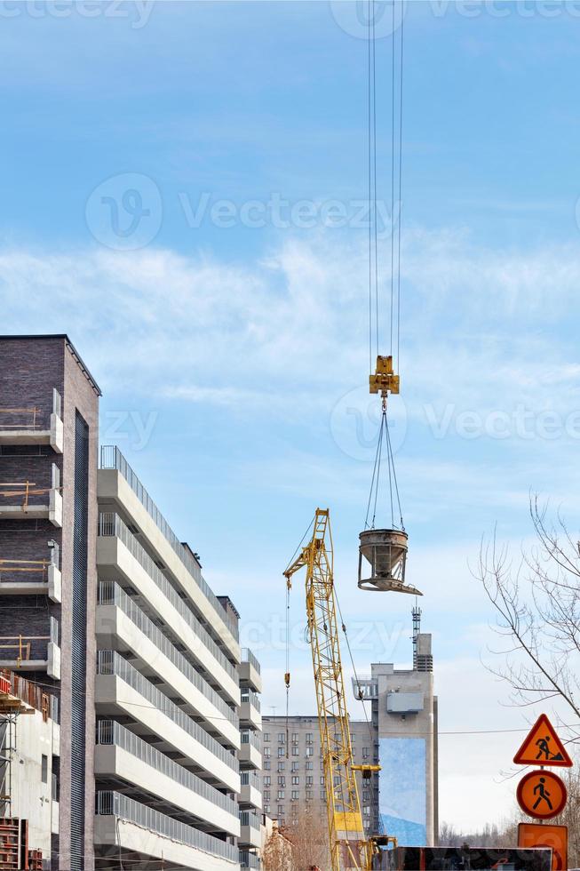 Concrete solution in a metal hopper is delivered to the construction site with a crane against a blue sky. photo