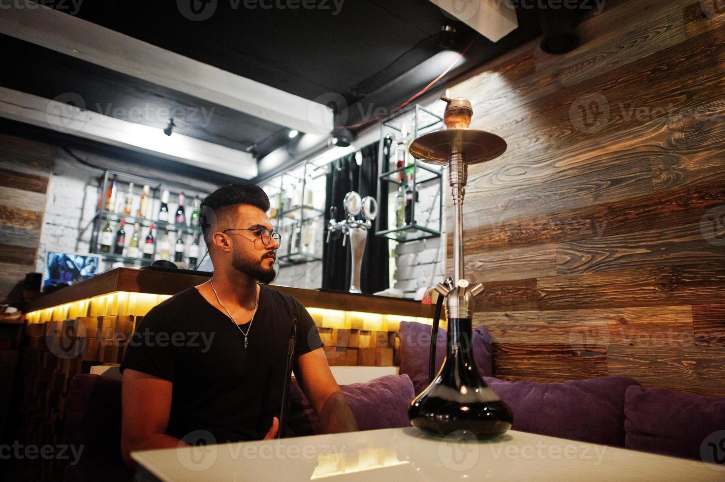 Stylish beard arabian man in glasses and black t-shirt smoking hookah indoor bar. Arab model having rest. photo